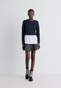 Pull marin avec un panneau inférieur blanc, associé à une jupe évasée grise et des bottes noires à lacets blancs. Design simple et moderne.
