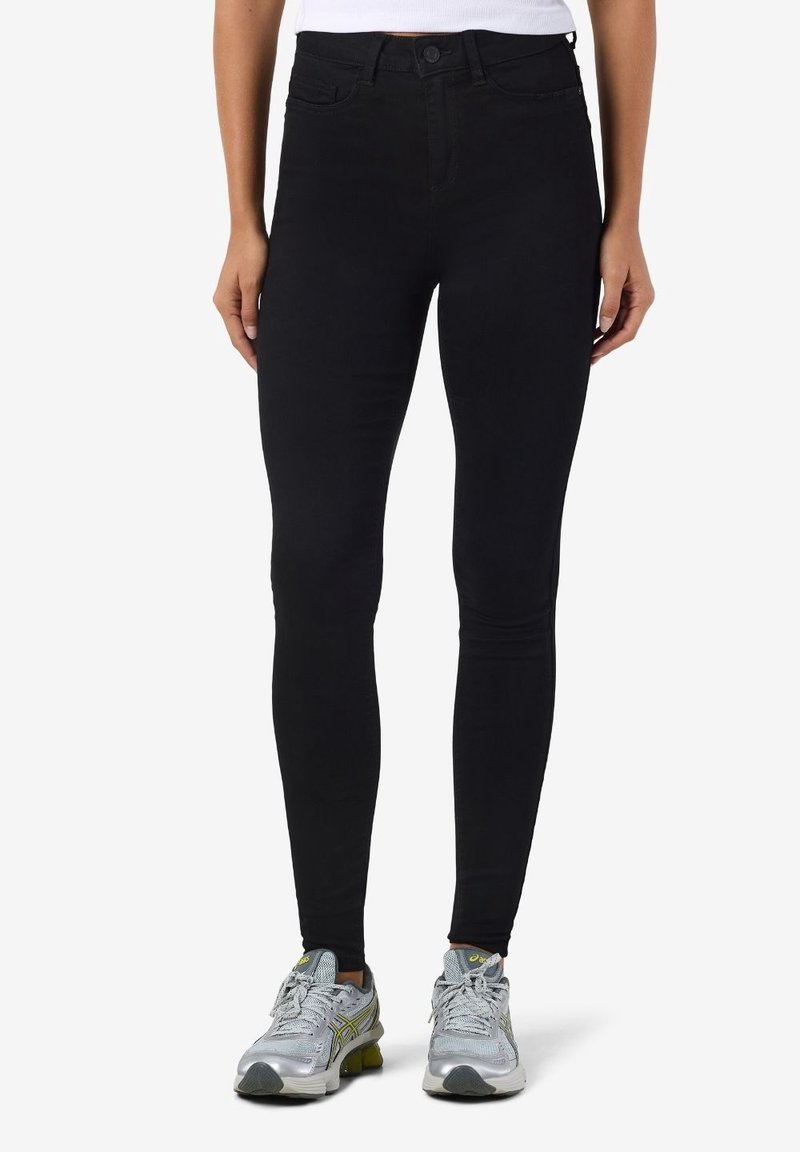 Femme portant un jean skinny taille haute noir et des chaussures de sport gris argenté, debout devant un fond blanc uni.