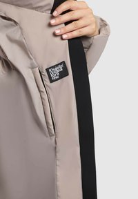 Hand die een open beige jasje met zwarte accenten vasthoudt, met zichtbare binnenzak en een zwart rechthoekig label met witte tekst.