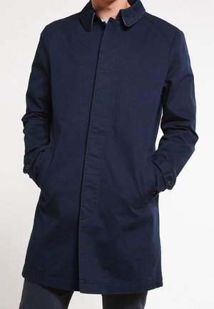 Mężczyzna w granatowym, długim płaszczu z kołnierzem, z rękami w kieszeniach, na jednolitym białym tle.