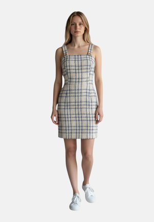 Giovane donna che indossa un vestito senza maniche, a quadri, lunghezza al ginocchio, con spalline larghe e sneakers bianche, in piedi di fronte su uno sfondo semplice.