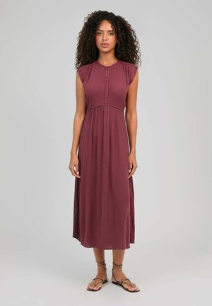 Femme aux cheveux bouclés porte une robe midi bordeaux sans manches et des sandales marron à lacets, debout devant un fond gris uni.