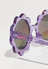 Ochelari de soare în formă de floare violetă, cu rame transparente strălucitoare și detalii cu logo roz pe picior. Lentilele reflectante reflectă lumina.