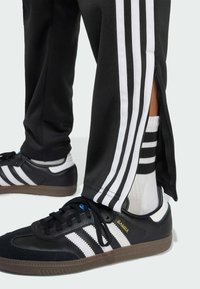 Zwarte sportbroek met witte zijstrepen en ritsmanchetten, gecombineerd met zwarte schoenen met witte strepen en een gestructureerde zool.