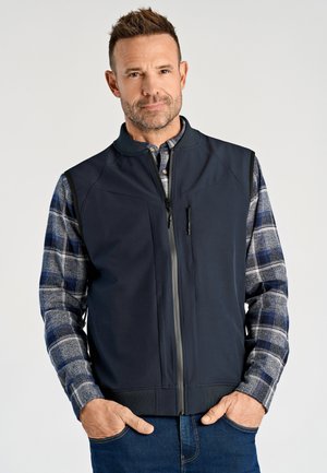 Mann trägt eine marineblaue Weste mit Reißverschluss über einem blau-grau karierten Hemd, hat die Hände in den Jeanstaschen und blickt nach vorne vor einem schlichten Hintergrund.