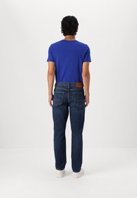 Blaues kurzärmliges Hemd kombiniert mit dunkelblauen Denim-Jeans. Die Jeans haben einen braunen Lederpatch am Bund und doppelte Gesäßtaschen.