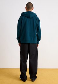 Teal huvtröja med avslappnad passform, utrustad med en känguruficka och ribbade muddarna, tillsammans med lösa svarta byxor på ett gult golv.