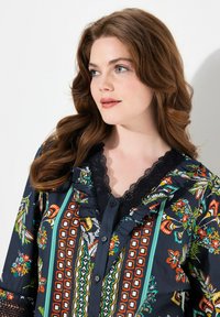 Camicia abbottonata con uno sfondo blu navy scuro, motivi floreali e geometrici in colori vivaci, dettagli in pizzo attorno al collo e bordi con volant.