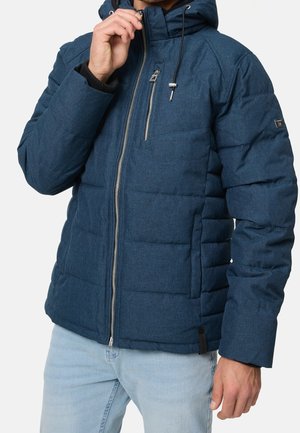 Homme portant une veste matelassée bleue avec capuche et fermeture éclair à l'avant, tirant la fermeture vers son menton, associée à un jean bleu clair.