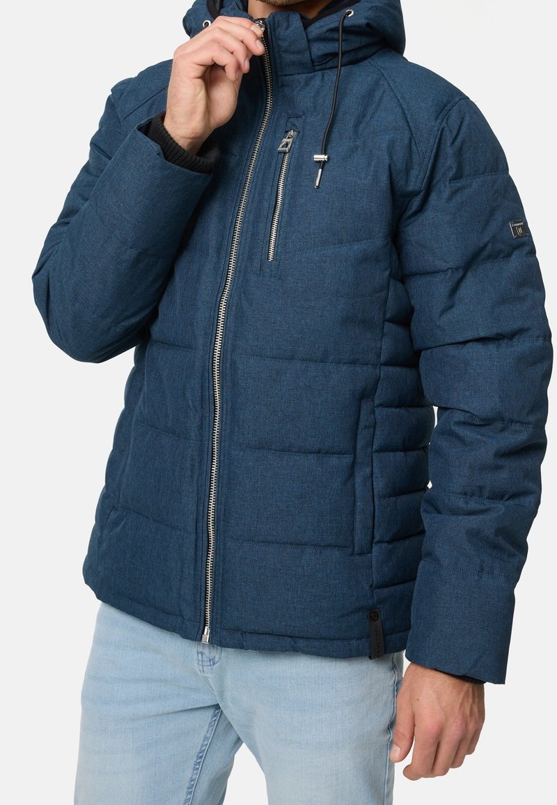 Homme portant une veste matelassée bleue avec capuche et fermeture éclair à l'avant, tirant la fermeture vers son menton, associée à un jean bleu clair.