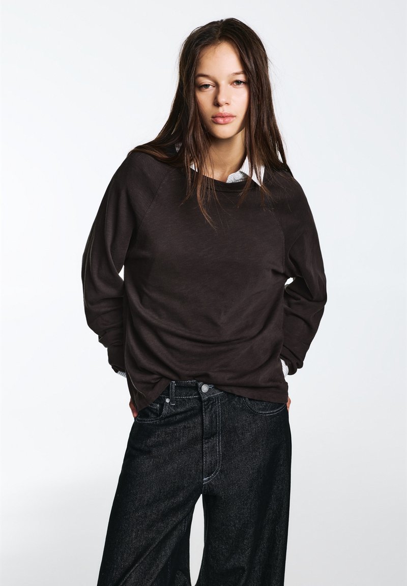 Camicia marrone scuro a maniche lunghe con scollo rotondo, abbinata a jeans denim neri a vita alta. La camicia presenta un colletto bianco a strati.