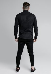 Schwarzes Langarmshirt mit tailliertem Schnitt, getragen zu schwarzen Slim-Jeans und schwarzen Schuhen. Verfügt über einen geknöpften Kragen und strukturierte Stoffoptik.