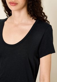 Moteris su garbanotais tamsiais plaukais, vilkinti juodą trumpomis rankovėmis, iškirptės formos marškinėlį ant šviesaus fono.