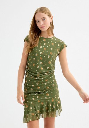 Joven mujer con un vestido verde ajustado de flores, mangas cortas y bajo con volantes, posando contra un fondo blanco liso.