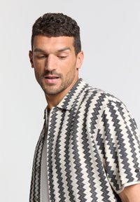 Korte mouw shirt met een zwart en crème zigzagpatroon, geweven textuur en een kraag. Voorzien van knopen aan de voorkant.