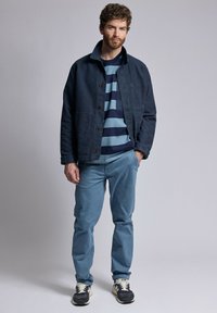 Veste en denim bleu avec un devant à boutons, superposée sur un t-shirt à col rond rayé bleu marine et bleu clair, accompagnée de pantalon bleu clair et de baskets.