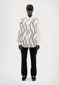 Camisa blanca de manga larga con un patrón de olas negras y texto, combinada con pantalones oscuros y zapatillas negras, llevada con una gorra negra.