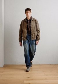 Brun bomberjakke, mørk skjorte, falmede blå jeans og grå sneakers. Modell som går på tregulv mot en nøytral vegg.