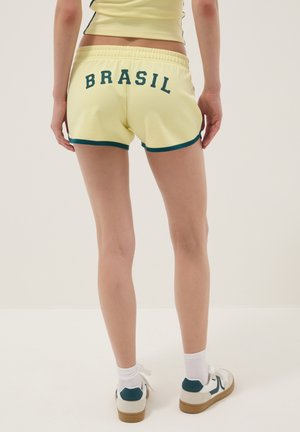 Persona que lleva pantalones cortos amarillos con el texto "BRASIL" y ribete verde, combinados con calcetines blancos y zapatillas blancas con detalles verdes.