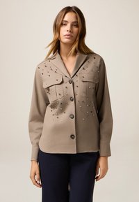 Beige Hemd mit Knöpfen und zwei Brusttaschen, verziert mit verstreuten metallischen Verzierungen und langen Ärmeln. Glatte Stofftextur.