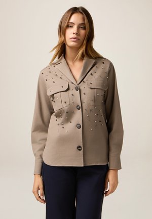 Beige Hemd mit Knöpfen und zwei Brusttaschen, verziert mit verstreuten metallischen Verzierungen und langen Ärmeln. Glatte Stofftextur.