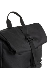 Mochila de tela negra con diseño de tapa enrollable, correa ajustable y cierre de clip. La textura es suave con detalles de costura visibles.