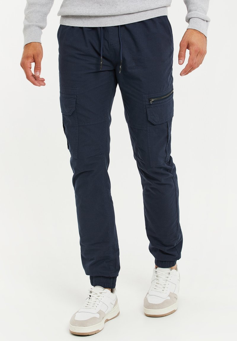 Pantalon cargo bleu marine avec ourlets élastiques, plusieurs poches dont une latérale zippée, et une taille à cordon de serrage. Associé à des baskets blanches et grises.