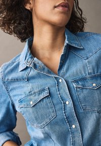 Jeansowa koszula w jasnoniebieskim kolorze, z klasycznym kołnierzykiem, zatrzaskami oraz dwiema przednimi kieszeniami z klapkami. Gładka tekstura i solidna konstrukcja.