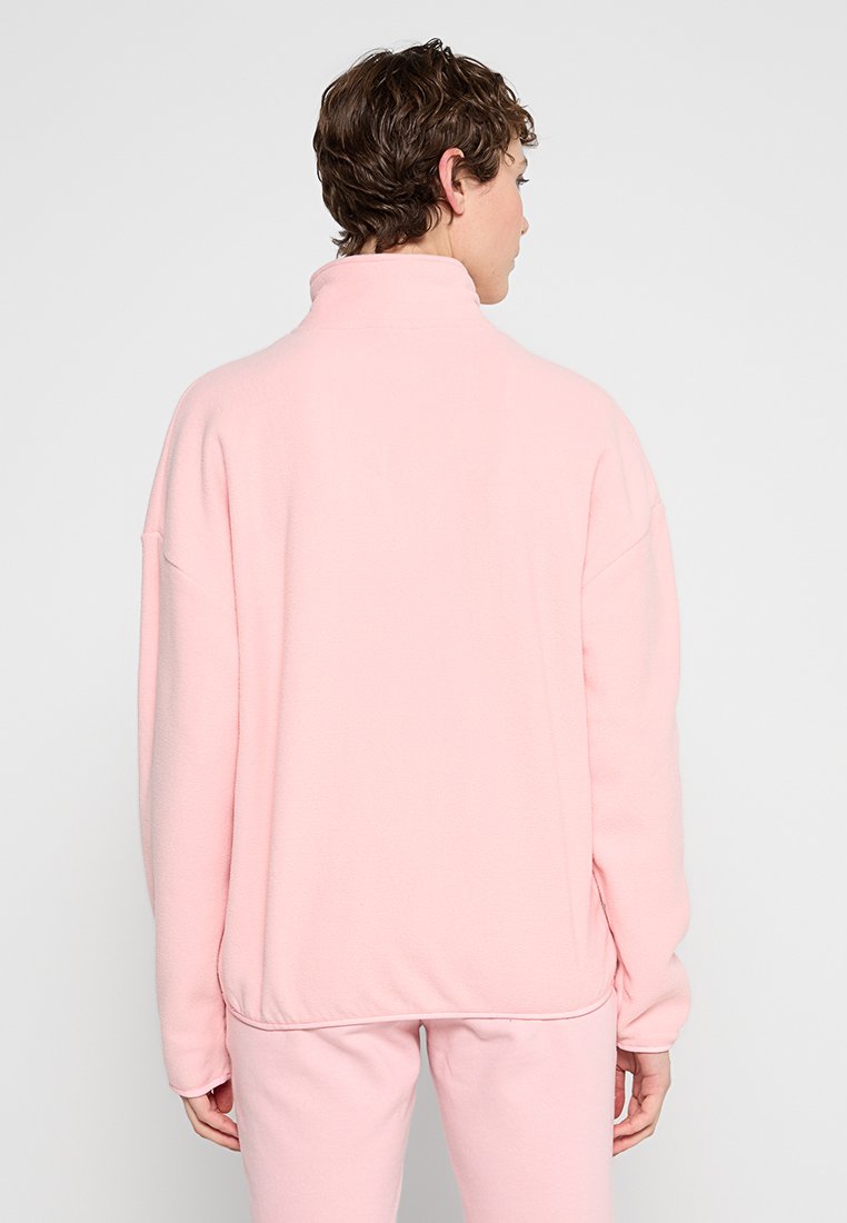 Pull en polaire rose avec un col montant, coupe décontractée et manches longues. Texture lisse, bas ourlé et détails de couture visibles aux épaules.