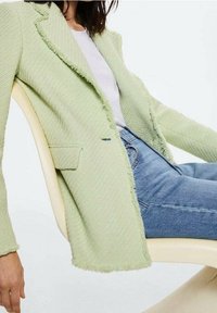 Jasnozielona marynarka o fakturowanej tkaninie z postrzępionymi krawędziami, pojedynczą kieszenią i klapami z wycięciem, noszona na białym t-shircie i niebieskich dżinsach.