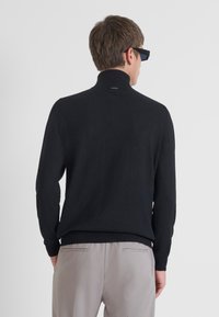 Maglione nero a collo alto in maglia con una texture liscia, caratterizzato da un colletto e polsini a coste, indossato sopra pantaloni grigio chiaro.