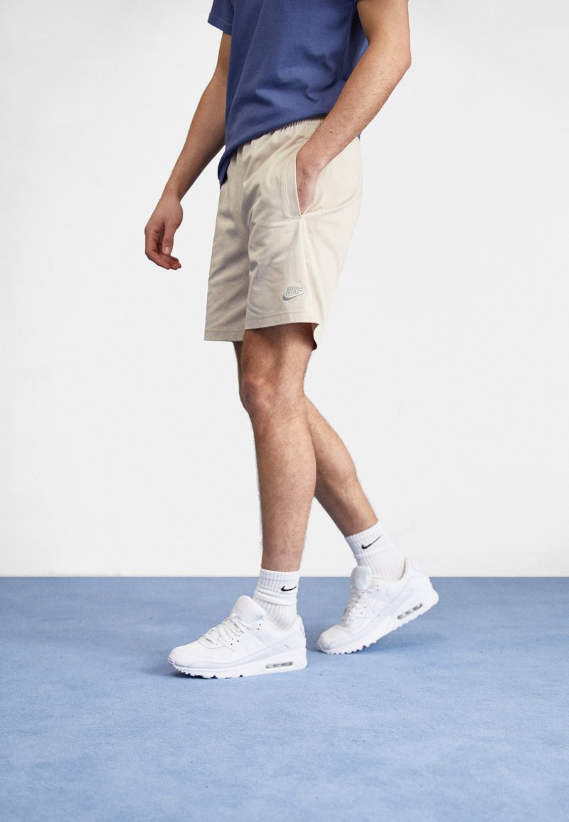 Beige athletic shorts with side pockets and a small Nike logo, paired with white sneakers and crew-length socks. Model in a blue t-shirt.