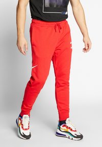 Röda svettbyxor med elastisk midja, sidofickor och vit Nike-logotyp. Matchas med flerfärgade träningsskor med djärva mönster.