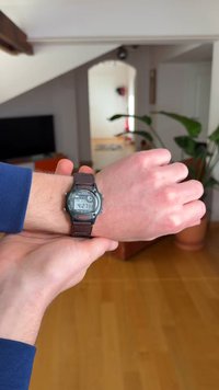 Digitale polshorloge met een zwarte kunststof behuizing, een bruin gestructureerde band en een LCD-display waarop tijd en datum worden weergegeven. Op de achtergrond staan kamerplanten.