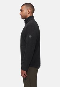 Giacca in pile nera con colletto alto, zip laterale e patch logo sul braccio sinistro. Tessuto morbido e vestibilità rilassata. Indossata con pantaloni verde oliva.