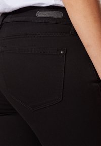 Jeans de mezclilla negra con un bolsillo trasero que presenta un sutil patrón de costura y una etiqueta de cuero en la cinturilla. Textura suave, ajuste entallado.