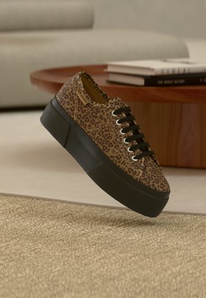 Sportiniai bateliai su leopardo raštu, juodu storu platforminiu padu ir juodomis raištelėmis, padėti ant kreminio kilimo ir medinio stalo su knygomis.