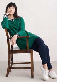 Groene tuniek met lange mouwen en een V-hals, met gestreepte mouwen, gecombineerd met donkere spijkerbroeken en witte sneakers, zittend op een houten stoel.