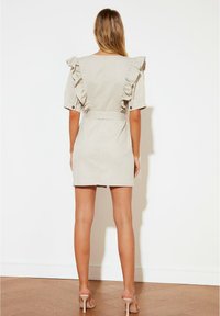 Béžové šaty s volánovými rukávy, upnutým pasem a rovný střih. Hladká textura, krátká délka, kombinované s neutrálními lodičkami na vysokém podpatku.