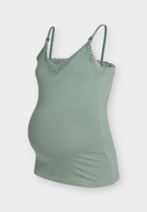 Femme enceinte en haut vert sans manches et pantalon court noir tenant une pochette noire, debout devant un fond blanc uni.