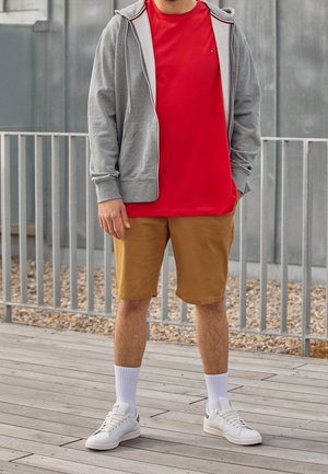 Mann steht im Freien und trägt ein rotes T-Shirt, einen grauen Kapuzenpullover mit Reißverschluss, beige Shorts, weiße Socken und weiße Turnschuhe auf einem Holzdeck.