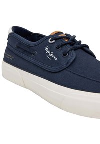 Pantof sport din pânză bleumarin cu talpă din cauciuc albă, având accente din piele maro, găuri rotunde pentru șireturi și logo-ul "Pepe Jeans London" pe lateral.