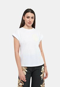 T-shirt bianca di cotone con maniche corte arrotolate, caratterizzata da un design floreale testurizzato sul lato sinistro del petto. Indossata con pantaloni scuri a fantasia floreale.