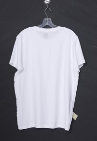 Hvid kortærmet T-shirt hængende på en klar plastik bøjle mod en mørk baggrund.