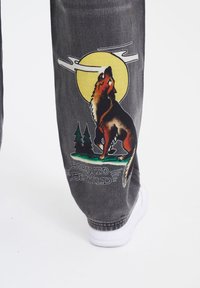 Grå denimjeans har et farverigt grafisk motiv af en ulv, der hyler mod en gul måne, accentueret med grønne træer og teksten "BORN TO BE WILD".