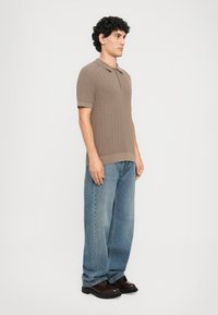 Mann med krøllete hår iført en brun kortermet zip-up strikket polo, vide blå jeans og mørkebrun lærsko, vendt mot høyre.