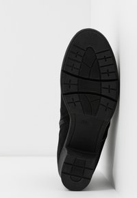 Semelle de botte noire avec motif de bande de roulement appuyée contre un mur d’angle blanc et un sol blanc.