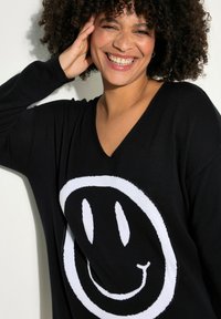 Maglione nero con una grande grafica di una faccina sorridente bianca al centro. Il tessuto sembra morbido con una vestibilità rilassata e design a V.