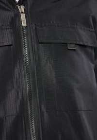 Sort jakke med frontlynlås, to øverste lommer og et subtilt tekstureret mønster. Stoffet virker let og sprødt.