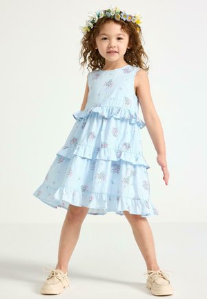 Ung pige iført en lyseblå blomsterkjole og blomsterkrans, stående med benene fra hinanden og armene let udstrakte mod en ensfarvet baggrund.